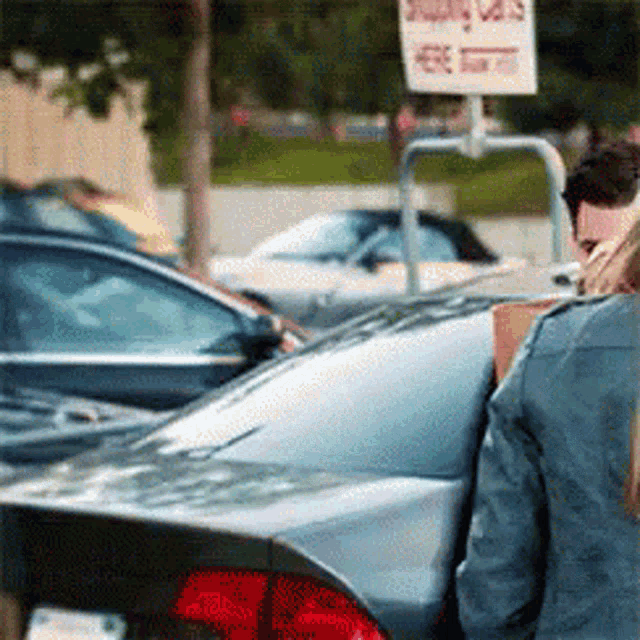 A man quickly ducks out of sight as a woman turns around, seemingly avoiding her or trying not to be noticed. The scene takes place in a parking lot next to a car, with a humorous sense of stealth or awkwardness as the man tries to hide.