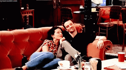 A man and woman sit cozily on a couch, with the woman resting her head on the man's shoulder. Both appear relaxed and happy, sharing a lighthearted moment in a warm, comfortable setting, likely a café. The scene evokes a feeling of closeness and affection between the two characters.