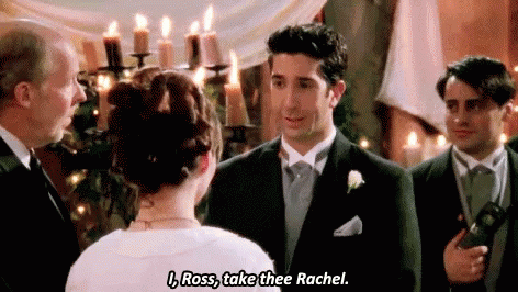 A man in a tuxedo, standing at the altar during a wedding ceremony, mistakenly says, "I, Ross, take thee Rachel," instead of the bride's name, causing a significant moment. The bride stands with her back to the camera, while a few people, including the officiant, react to the slip-up. The scene is lighthearted and awkward, highlighting a humorous and iconic wedding blunder from a famous TV show.