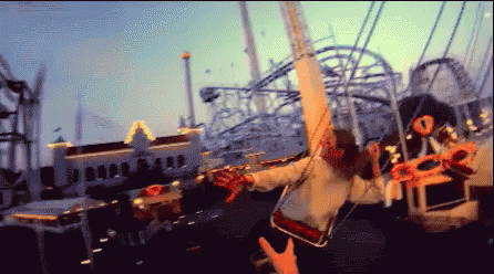 The image shows a person on a carnival swing ride, with arms outstretched and the ride in full motion. The background features other amusement park attractions, including roller coasters, illuminated by the evening light. The perspective captures the excitement and thrill of the ride, with the motion blur adding a dynamic, fast-paced feel to the scene.