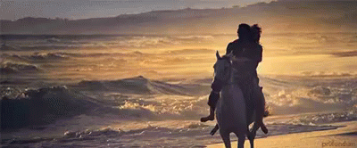 The image shows a romantic scene of a couple riding a horse along the beach at sunset. The sun casts a golden glow over the ocean waves and the sandy shore, creating a serene and picturesque atmosphere. The couple is seated close together, holding onto each other as they ride, capturing a peaceful and intimate moment by the sea.