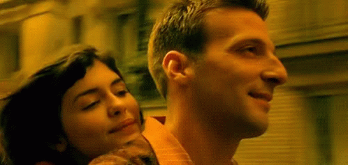 <br>A woman is resting her head peacefully on the back of a man's shoulder as they ride together on a motorcycle or scooter through a city. The scene has a warm, golden hue, giving it a nostalgic and romantic feel, suggesting a moment of connection and happiness between the two characters.