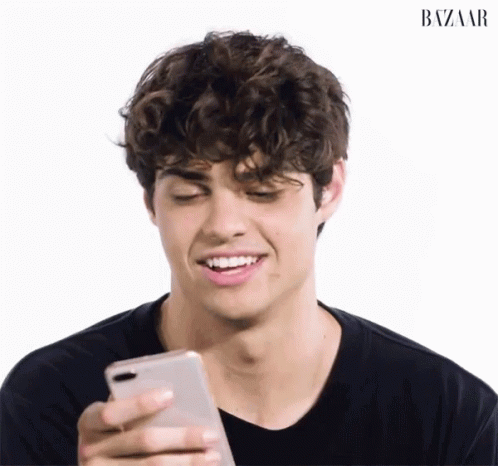 A young man with curly hair smiles as he looks down at his phone, appearing amused. The image is set against a plain white background, with "BAZAAR" written in the top right corner.