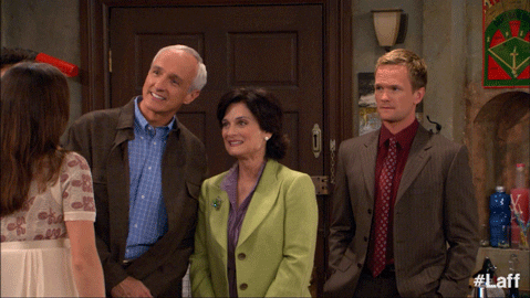 A group of three people, two older adults smiling warmly, and a younger man standing beside them looking slightly uncomfortable or awkward. They are indoors, possibly in a family setting, and appear to be greeting someone at the door.