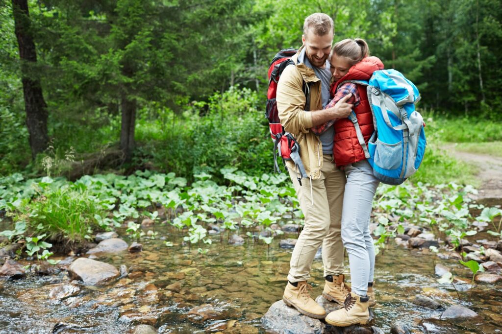 The image shows a couple hiking in a forest, standing close together on rocks by a shallow stream. Both are wearing outdoor gear, including backpacks and hiking boots. The woman is snuggled into the man’s chest, and they are smiling, enjoying a tender moment in nature. The lush greenery of trees and plants surrounds them, creating a peaceful and adventurous atmosphere.