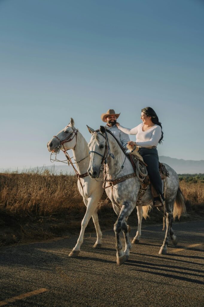 The image shows a couple riding side by side on two white horses through a rural landscape. The man, wearing a cowboy hat, looks at the woman with a smile, while she, dressed in a white top, reaches over playfully toward him. The setting is calm and scenic, with open fields and mountains in the distance under a clear blue sky, giving the moment a relaxed and romantic atmosphere.