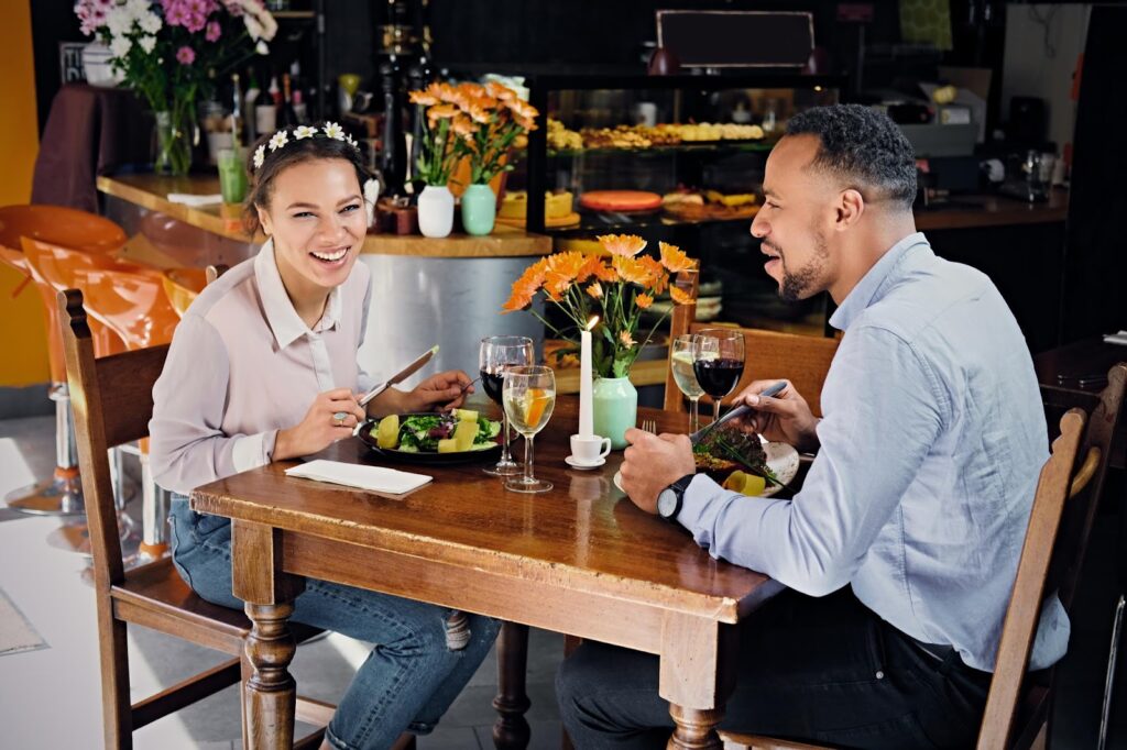 The image shows a couple sitting at a cozy restaurant table, enjoying a meal together. The woman is smiling brightly while holding a fork and knife, with a salad in front of her. The man is mid-conversation, holding a fork, with a plate of food and a glass of wine. A candle and a vase of flowers are on the table, adding a warm and romantic touch to the setting. The restaurant atmosphere is inviting, with soft lighting and a display of pastries in the background.