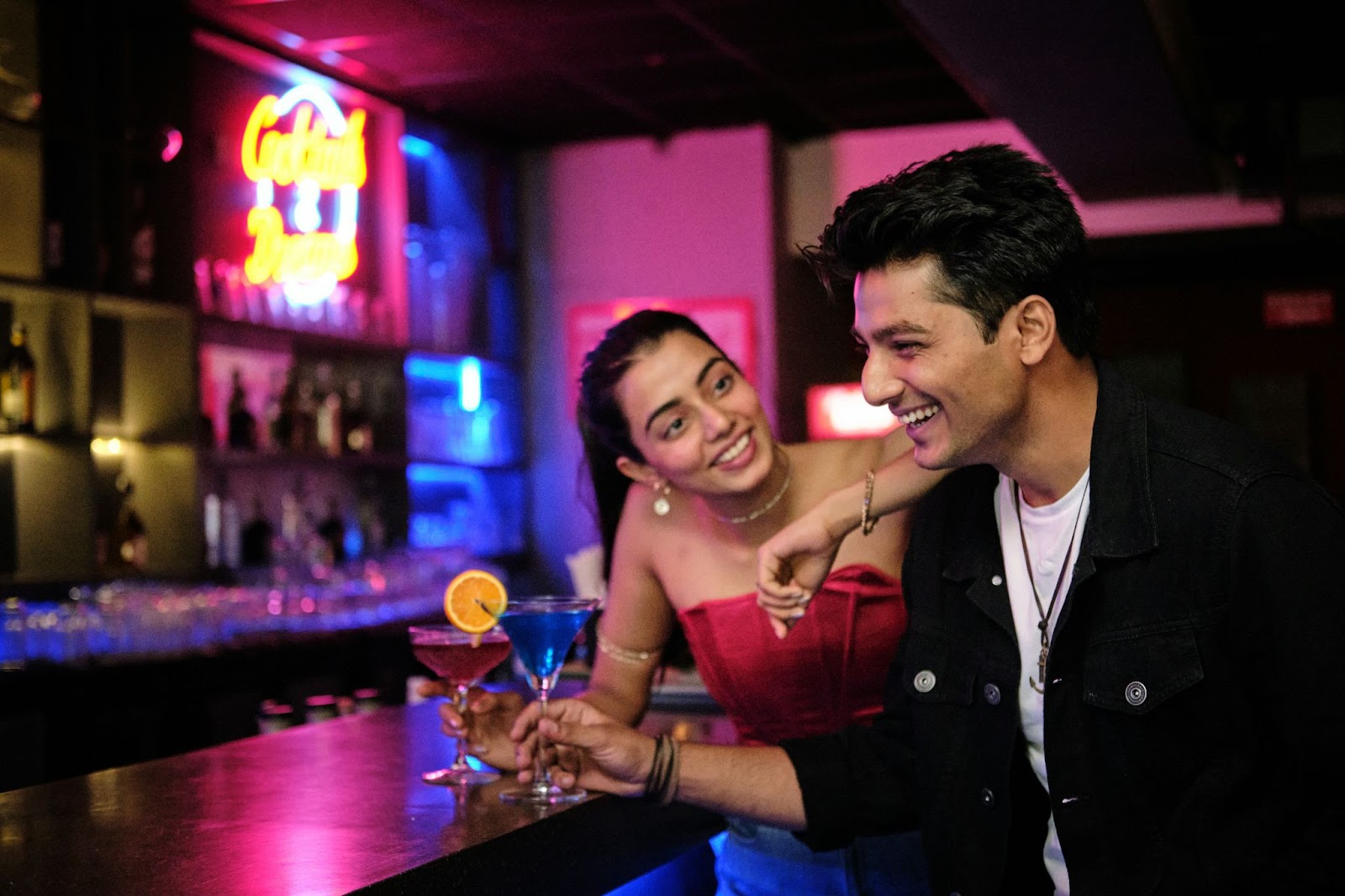 A smiling man and woman are enjoying drinks at a bar, with colorful neon lights glowing in the background. The woman leans playfully on the man's shoulder while they share a fun, lively moment. Both have cocktails in front of them, adding to the relaxed, cheerful atmosphere.