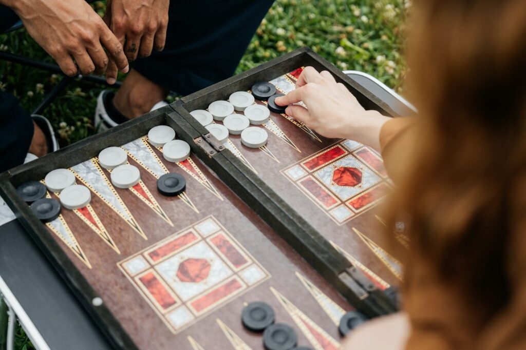 The image shows two people playing a game of backgammon outdoors. One person is moving a black game piece, while the other’s hands rest nearby, waiting for their turn. The board has an intricate design with red and white patterns, and the setting appears to be a casual, relaxing moment, likely in a park or backyard, as grass is visible in the background.