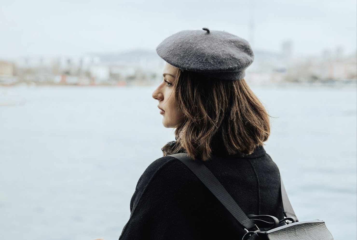 Woman wearing a gray beret and black coat, looking out over a body of water with a city in the distance.