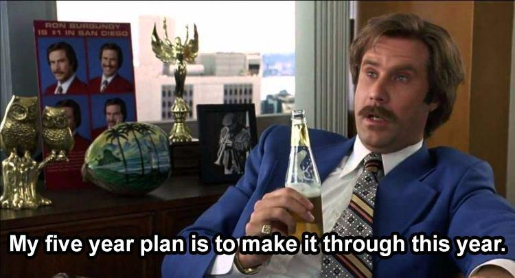 A man in a suit, holding a beer, sits casually in an office setting with the caption, "My five year plan is to make it through this year." The scene humorously conveys a sense of laid-back attitude and low expectations, with the character showing a nonchalant approach to future planning.